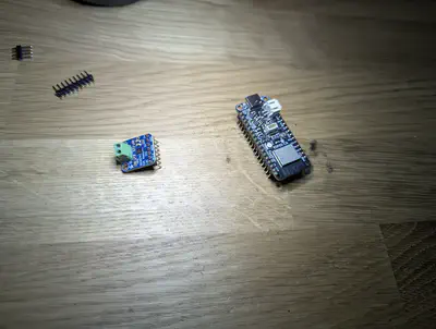 On a wooden desk, a small blue Adafruit MAX98357A I2S amplifier breakout with a green screw terminal on the left, and a larger black Adafruit ESP32-C6 Feather board on the right, with two loose header strips above them