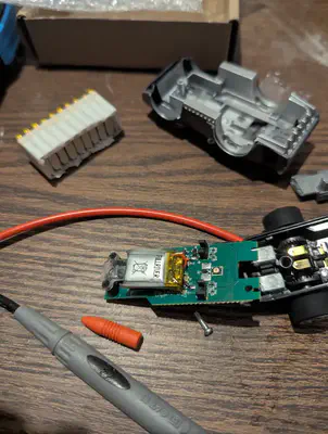 An opened Anki Overdrive car on the workbench, with its battery on the circuit board, exposed motors and gears, the removed body shell nearby and multimeter probes in the foreground.