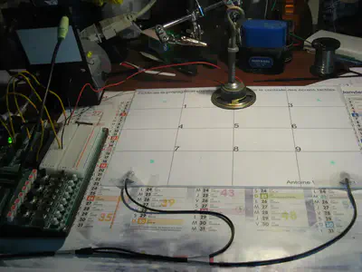 The 2009 experimental rig: a calendar page used as a 9-position grid, a small breadboard on the left, a speaker on a stand, two black microphone cables running to the bottom of the plate, with a handwritten title that reads &ldquo;Étude de la propagation d&rsquo;ondes dans le contexte des écrans tactiles&rdquo;